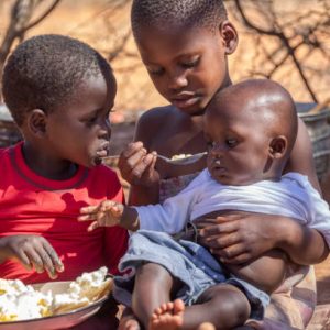 three african children in a village near Kalahari desert, the sister feeding her brother in the outdoors kitchen